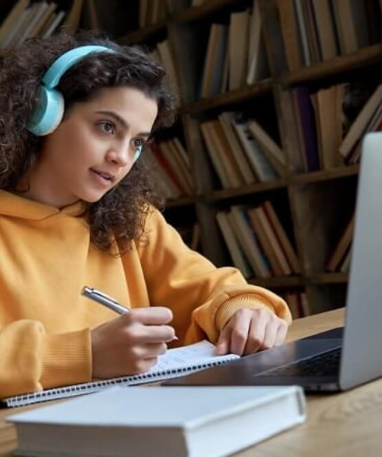 A young high school student wearing headphones and taking notes in front of her computer for an online internship meeting