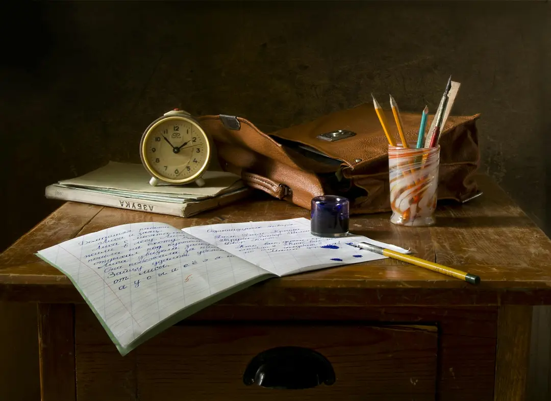 Open notebook lying on a desk, surrounded by a clock, pen pot and satchel